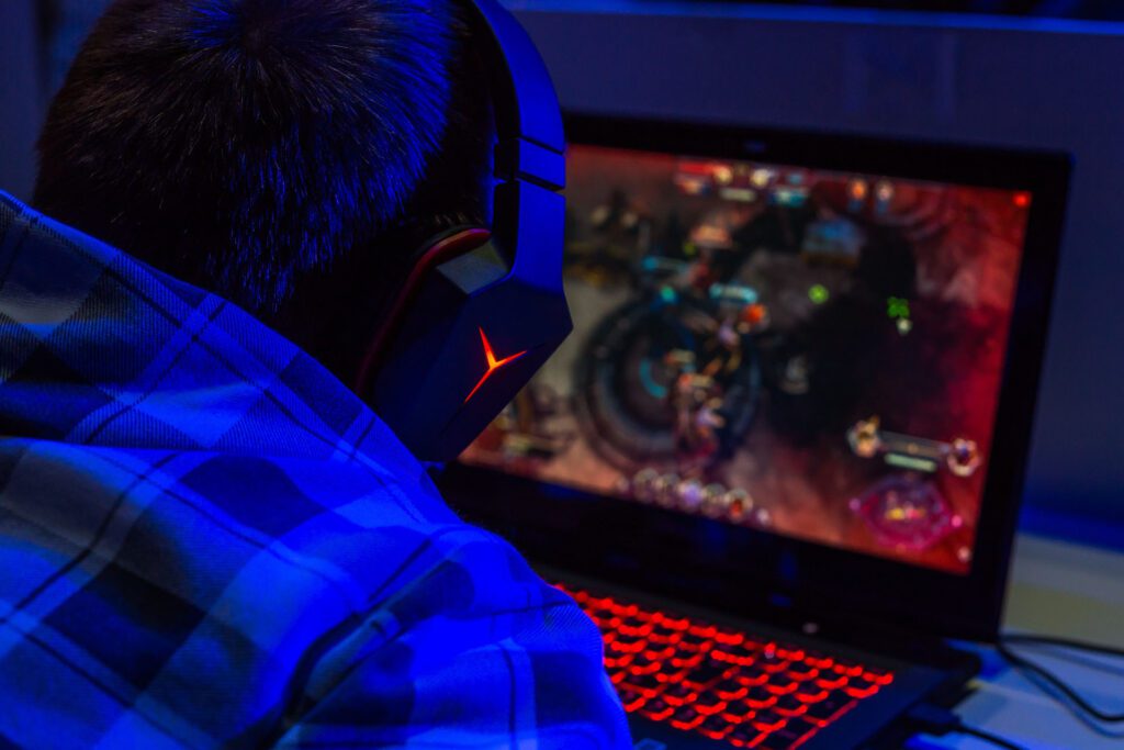 Man with headphones sitting down at his laptop playing a video game with an illuminated keyboard.