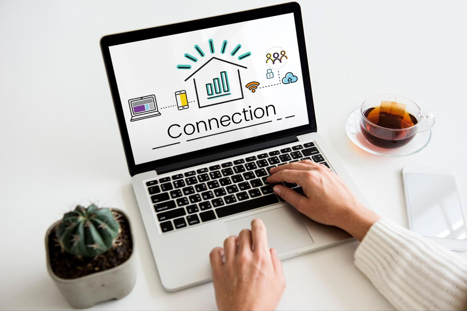 Woman wearing a sweater typing on a computer with the word “connection” on the screen with various icons next to a potted cactus, a cup of tea, and a cell phone.